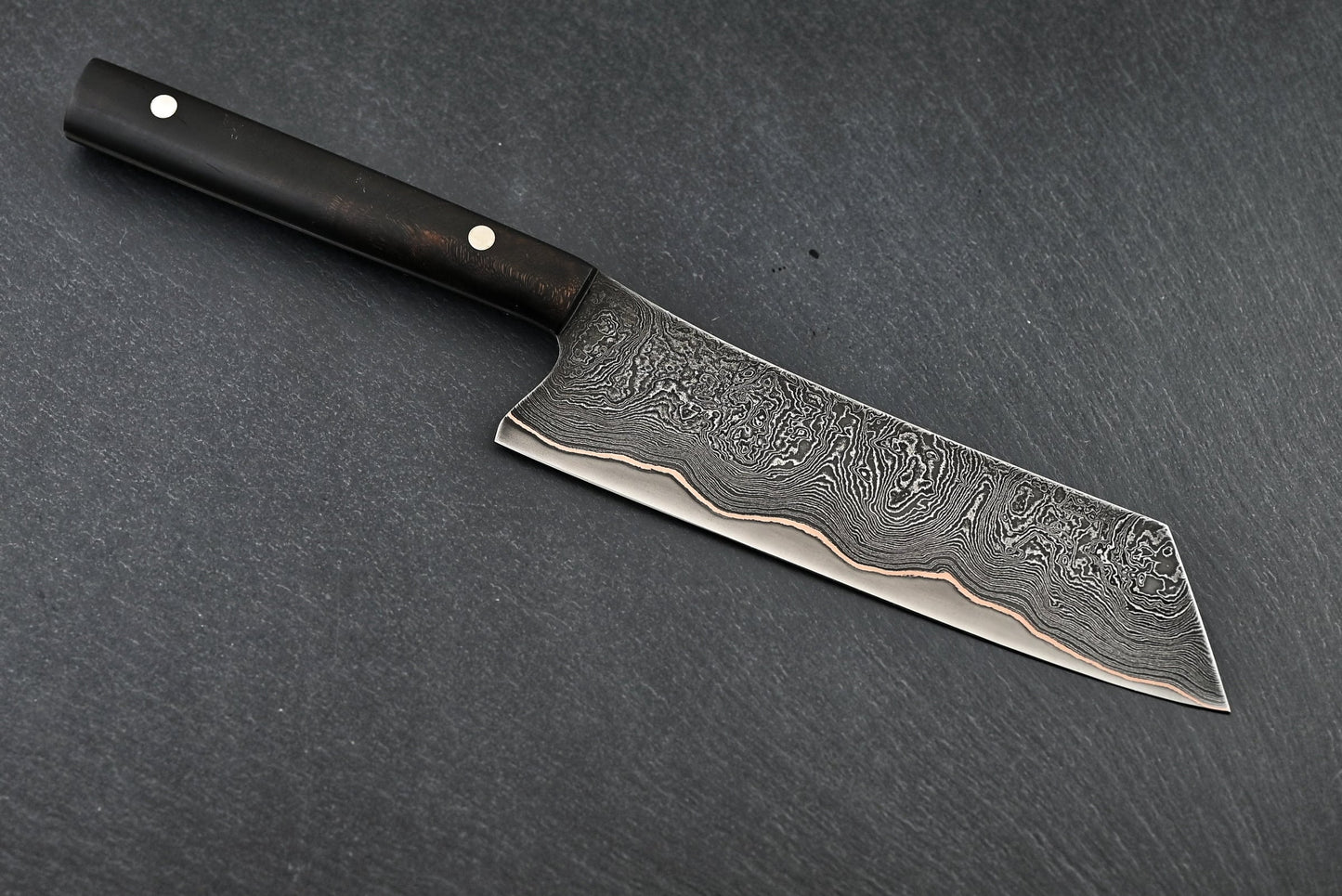 Santoku-Messer: Damastmuster-Klinge mit schwarzem Griff, schräg auf dunkler Fläche.